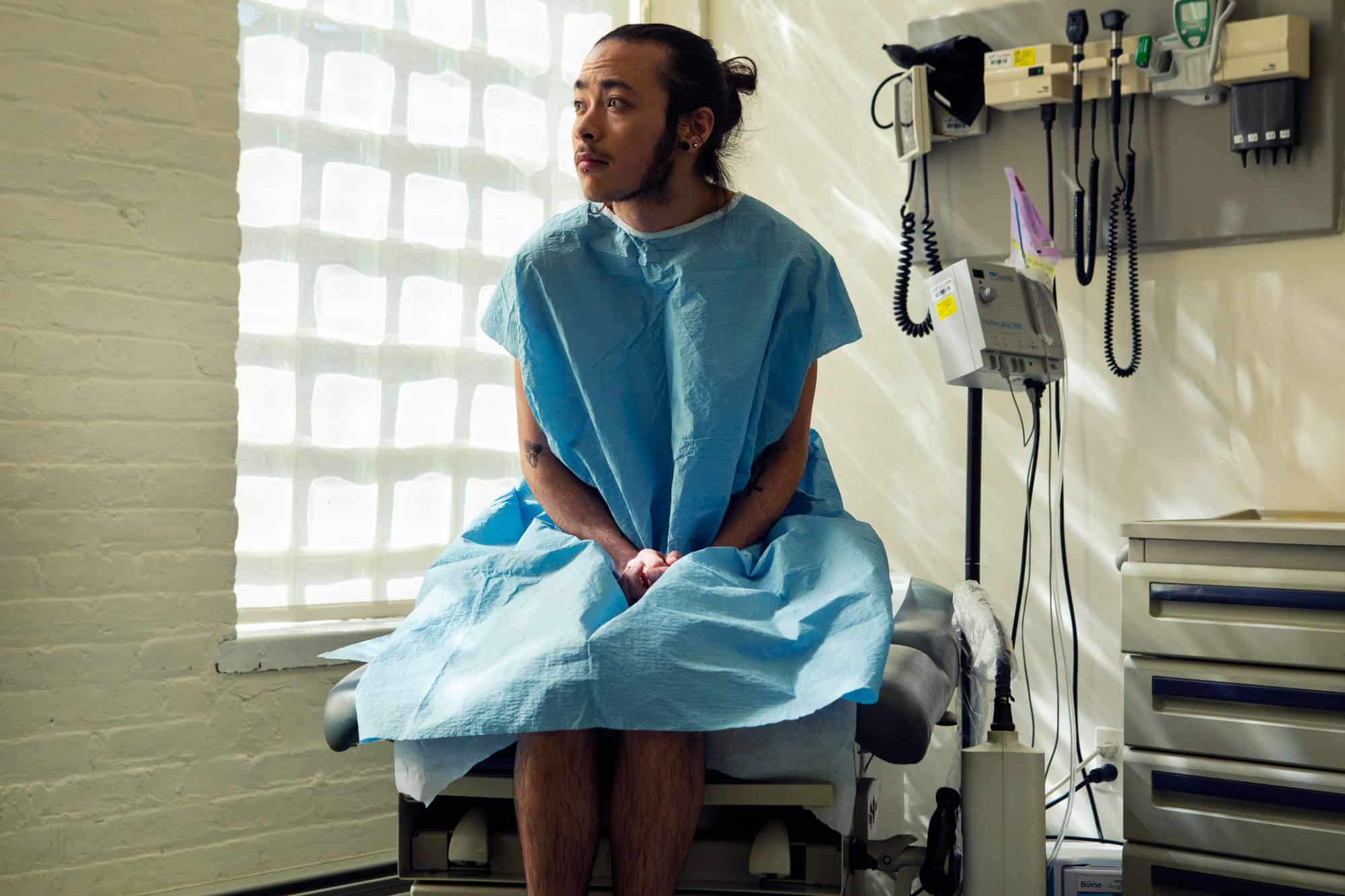 A genderqueer person sitting in a hospital gown in an exam room