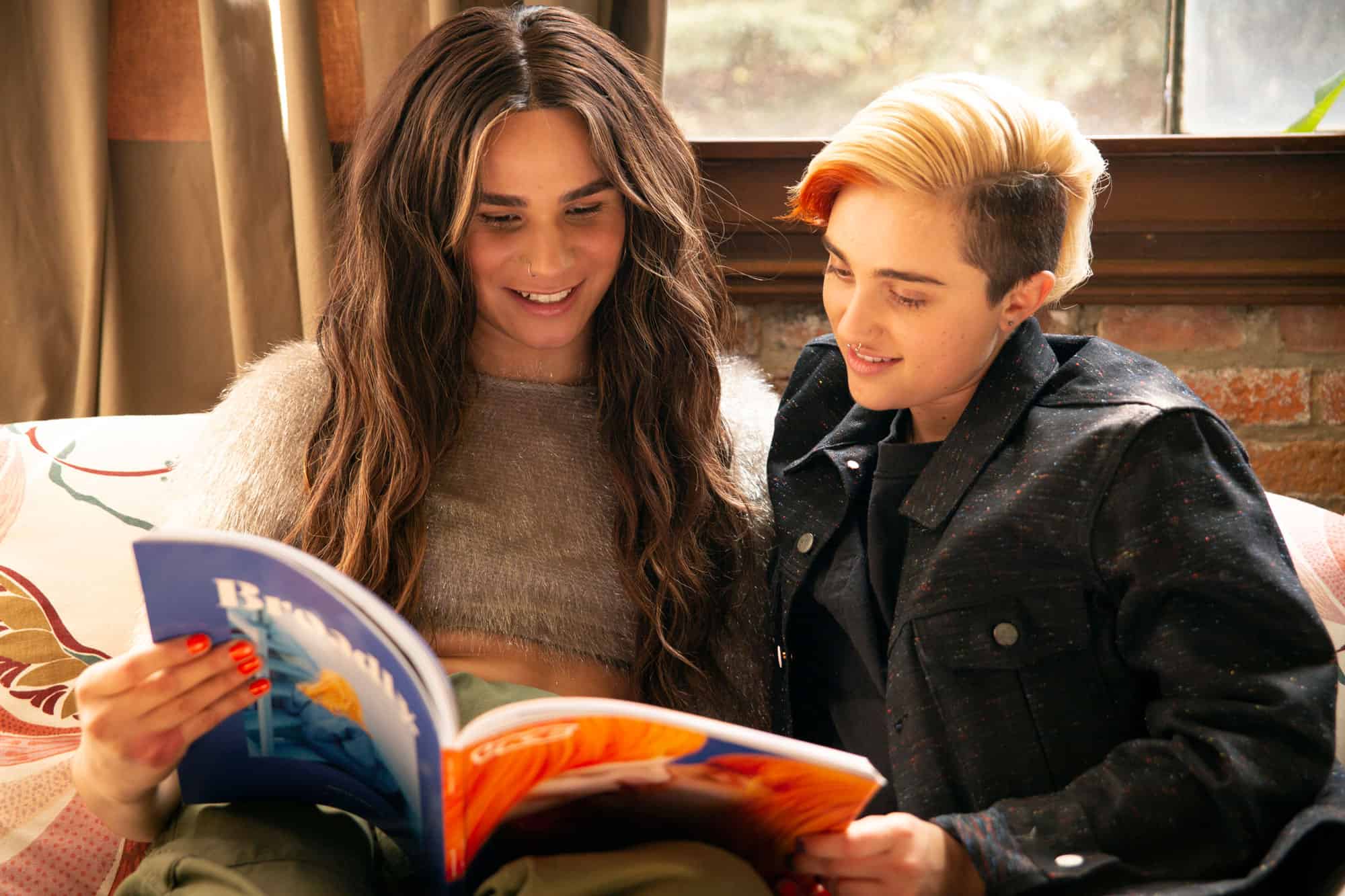 A transfeminine non-binary person and transmasculine gender nonconforming person reading a magazine together